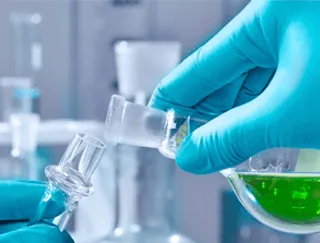 Laborant mit Handschuhen gießt grüne Flüssigkeit in Glasapparatur.